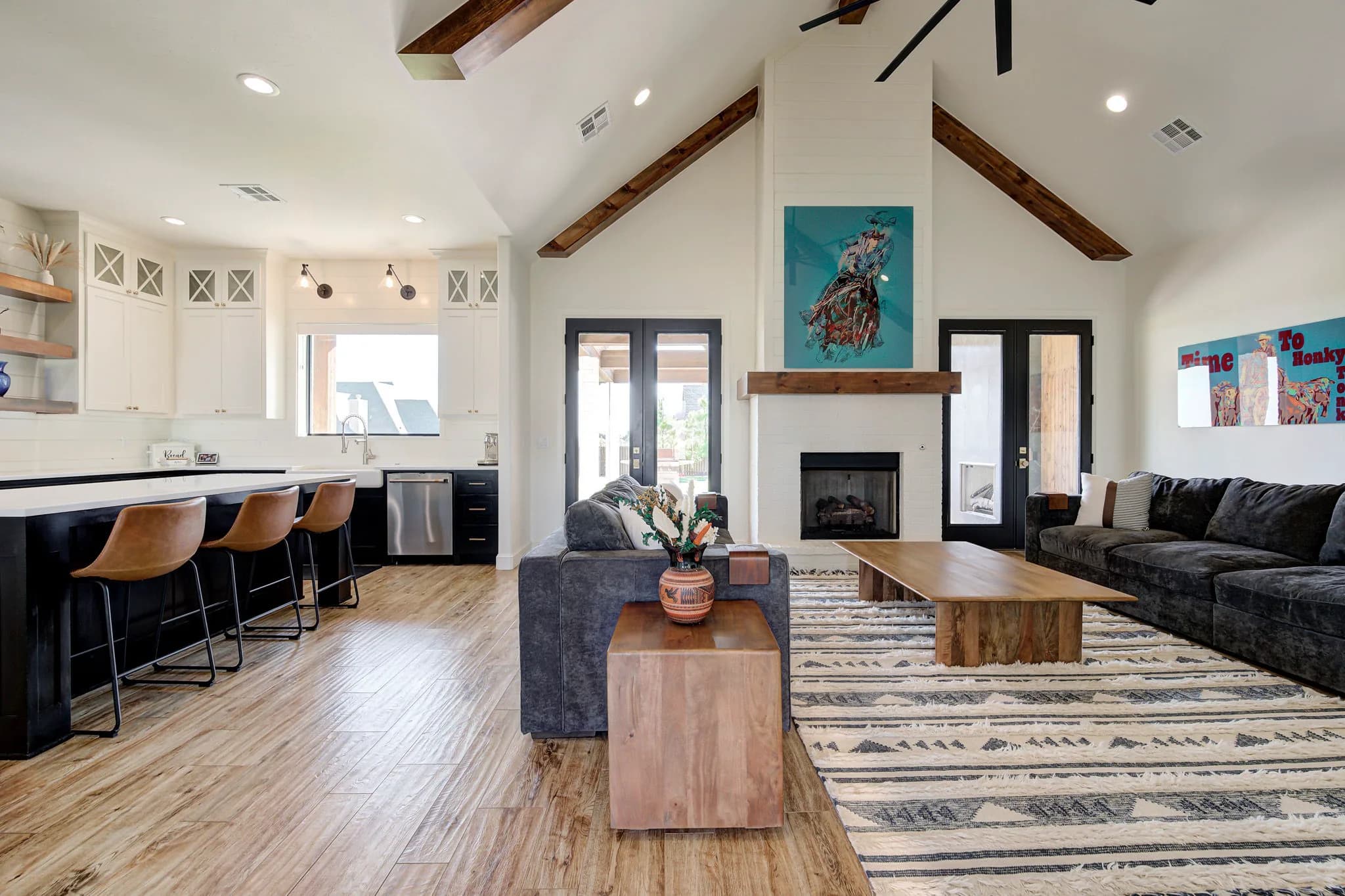 Vaulted great room with exposed wood beams, a white-brick gas fireplace, and an open kitchen beyond