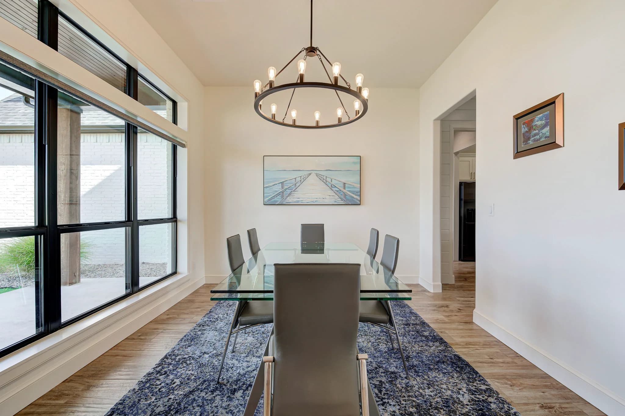 Formal dining room with a glass-top table, upholstered chairs, a round bulb chandelier, and black-framed windows overlooking a courtyard