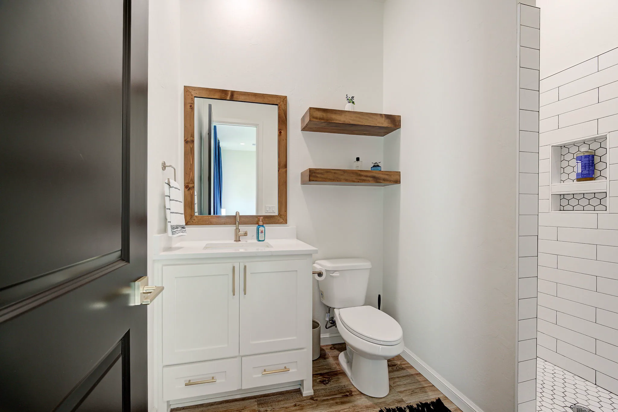 Guest bathroom with a white vanity, a wood-framed mirror, floating shelves, and a subway-tile walk-in shower