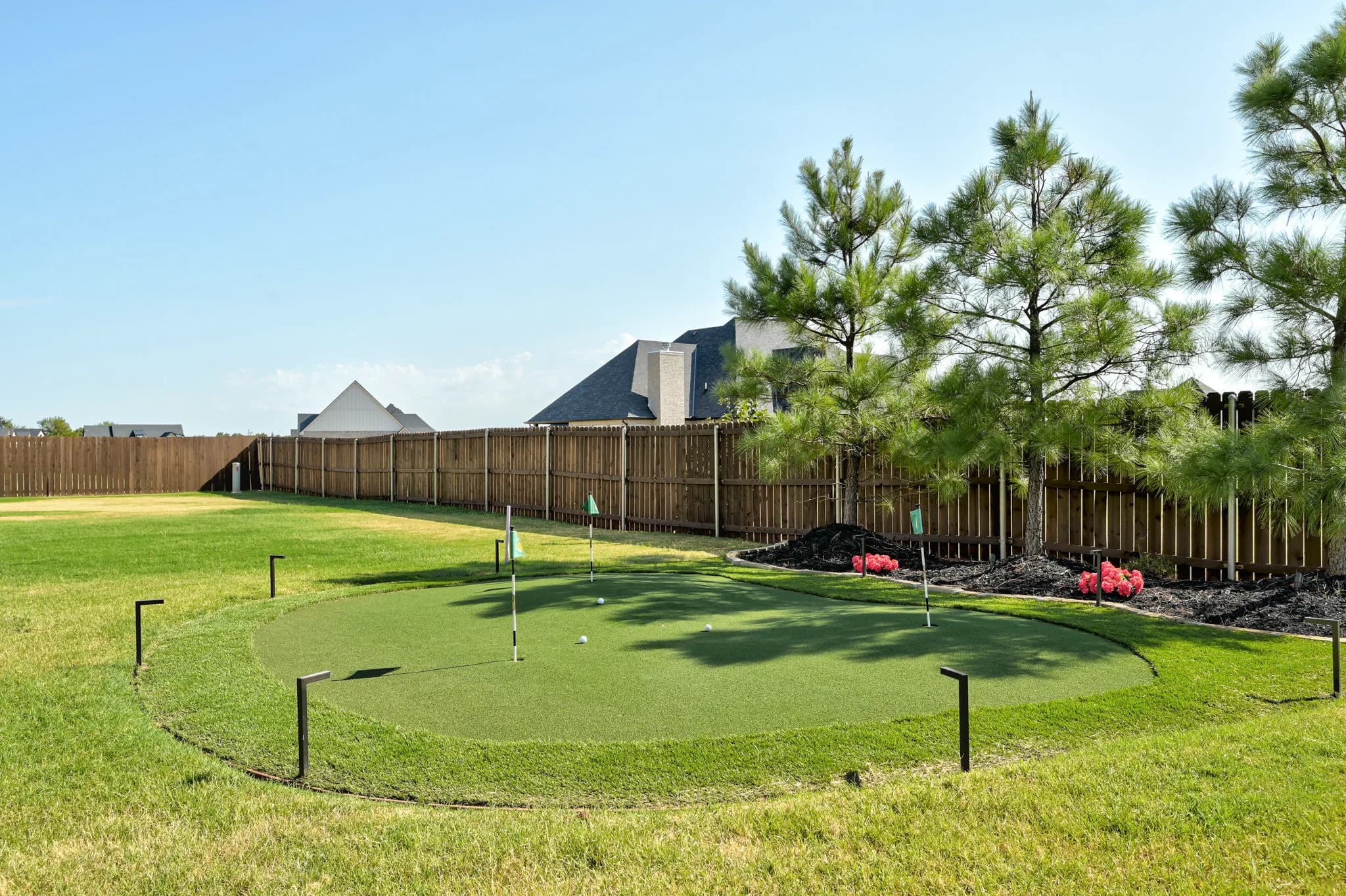 Backyard putting green with multiple flags, bordered by pine trees and landscaped beds along a privacy fence